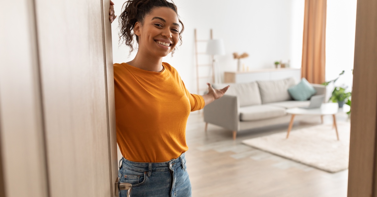 A young person in an orange top and jeans opening the door to their new home in a welcoming gesture.