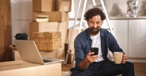 A person sitting in their new home and smiling at their phone. They are surrounded by cardboard moving boxes.