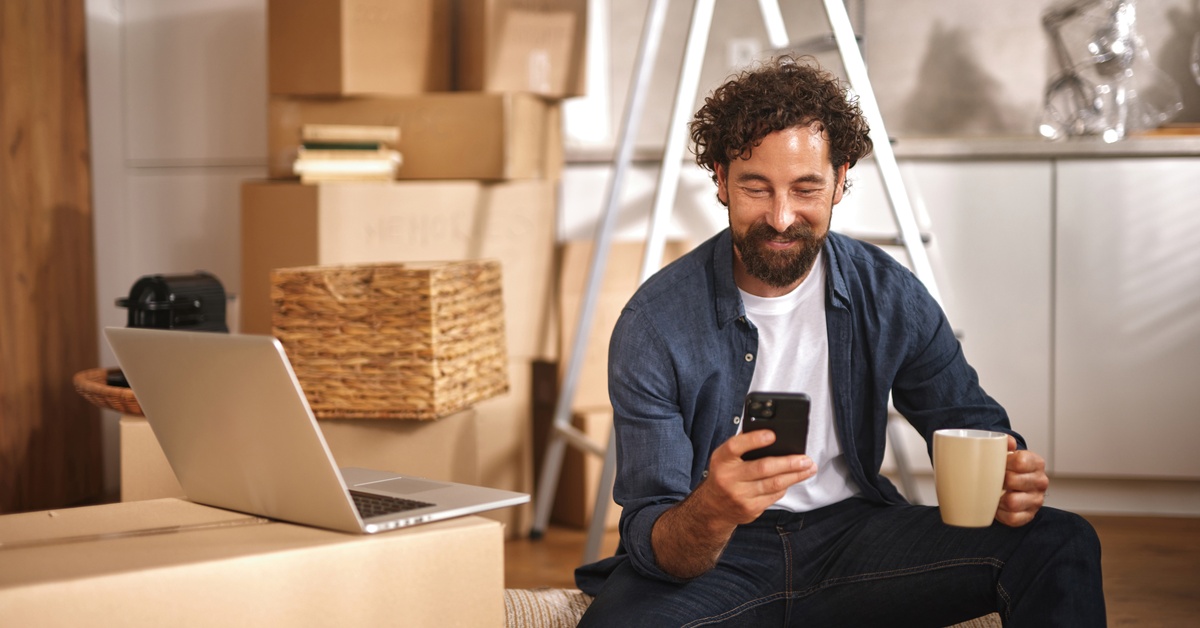 A person sitting in their new home and smiling at their phone. They are surrounded by cardboard moving boxes.