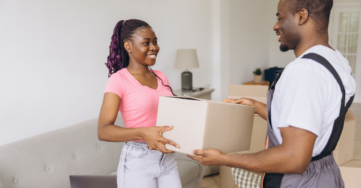 A moving professional handing off a small cardboard box to a customer who is standing in her new home.
