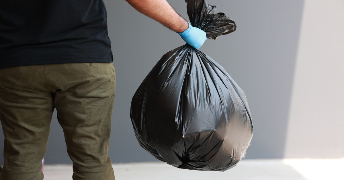 A person standing in an empty white space while wearing blue gloves and holding a full black trash bag.
