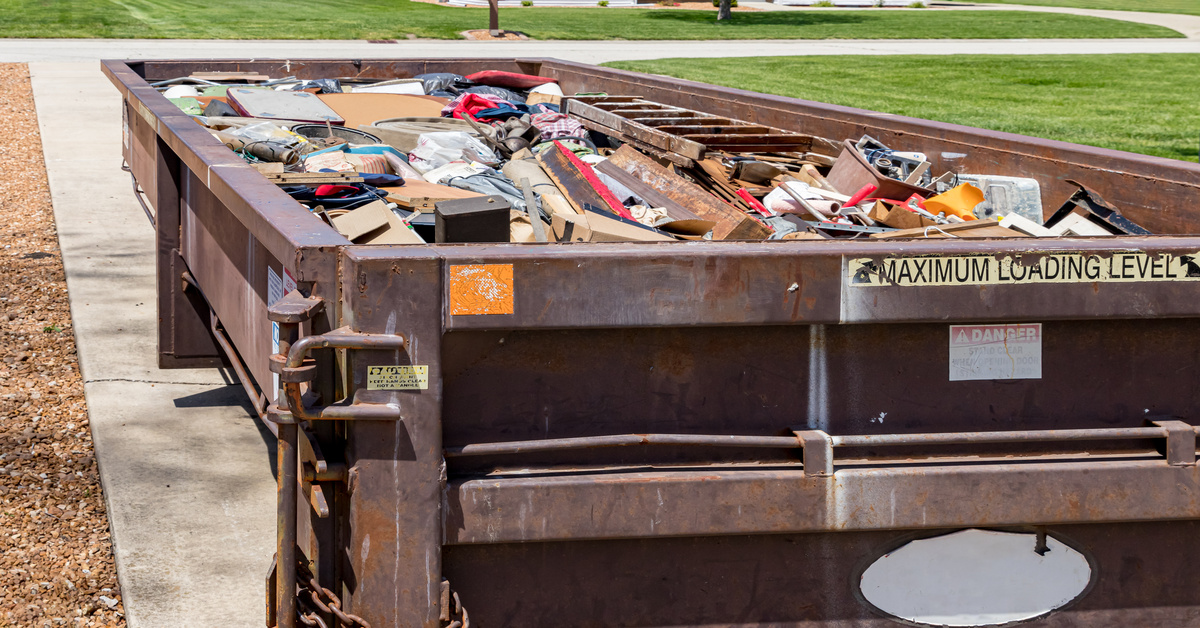 A large brown dumpster sitting in a residential driveway. It is full of different types of junk from a home.