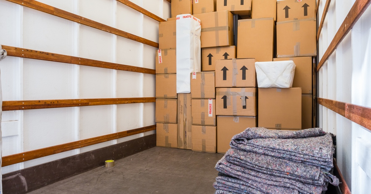 Moving boxes are stacked on top of one another in the back of a moving truck. In the foreground is a stack of furniture pads.