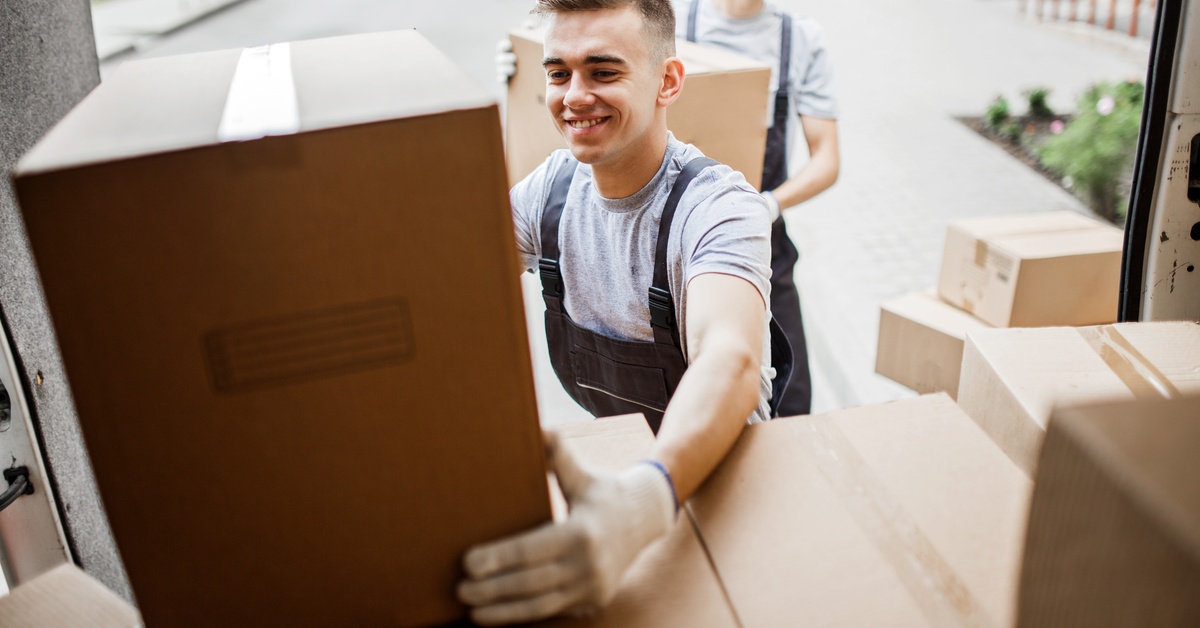 Two smiling young men lift moving boxes on top of other boxes in a moving truck.