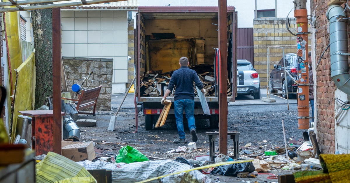 A professional in gloves carrying debris to the back of a moving truck. He is clearing out an alley full of small junk.