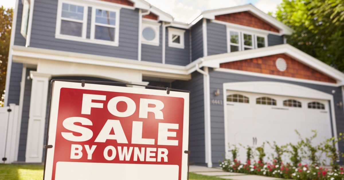 A red-and-white "For sale by owner" sign standing in the clean yard of a two-story blue, white, and brown home.