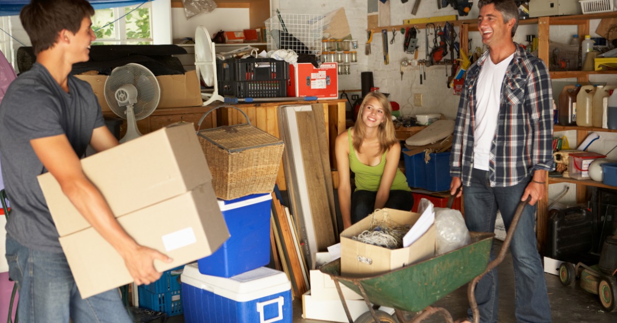 A group of smiling people moving items out of a cluttered garage in boxes and in a wheel barrow.