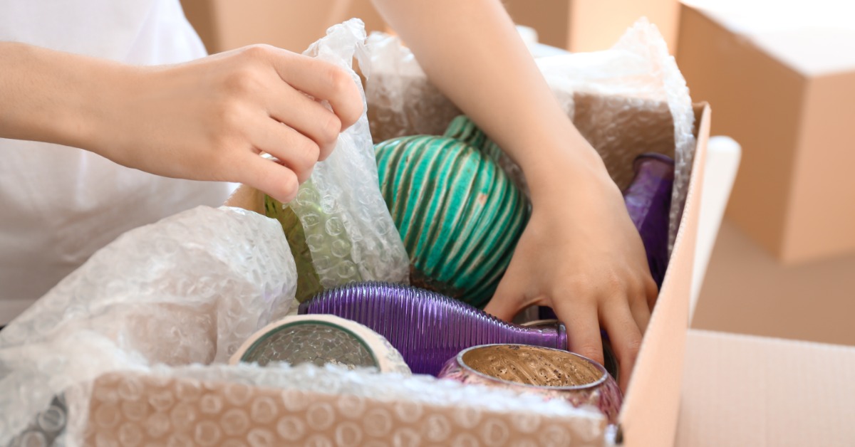 A person packing multiple fragile items, including vases and pottery, into a moving box with bubble wrap.